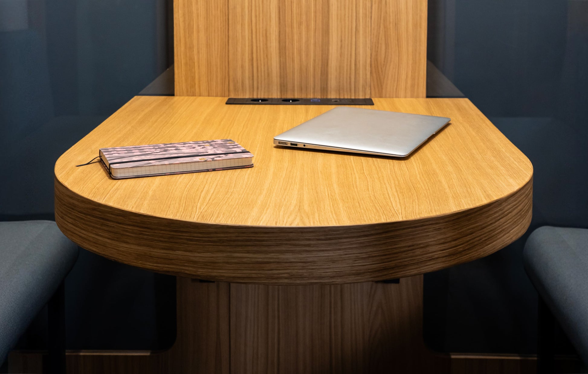 Haven Four meeting pod featuring a sleek oval oak table with a laptop and notebook, showcasing modern workspace design in warm wood tones against dark upholstered seating and walls, perfect for intimate 2-4 person collaborations