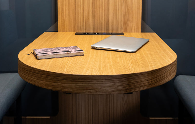 Haven Four meeting pod featuring a sleek oval oak table with a laptop and notebook, showcasing modern workspace design in warm wood tones against dark upholstered seating and walls, perfect for intimate 2-4 person collaborations